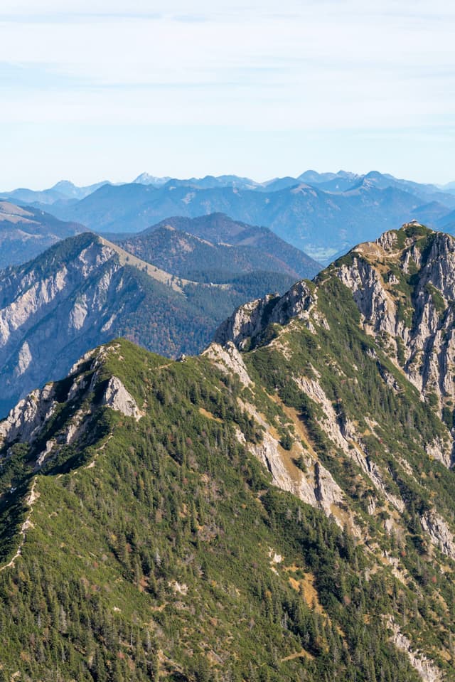 Jagged green-covered mountain ridge with steep rocky slopes and layers of distant blue peaks under a pale sky