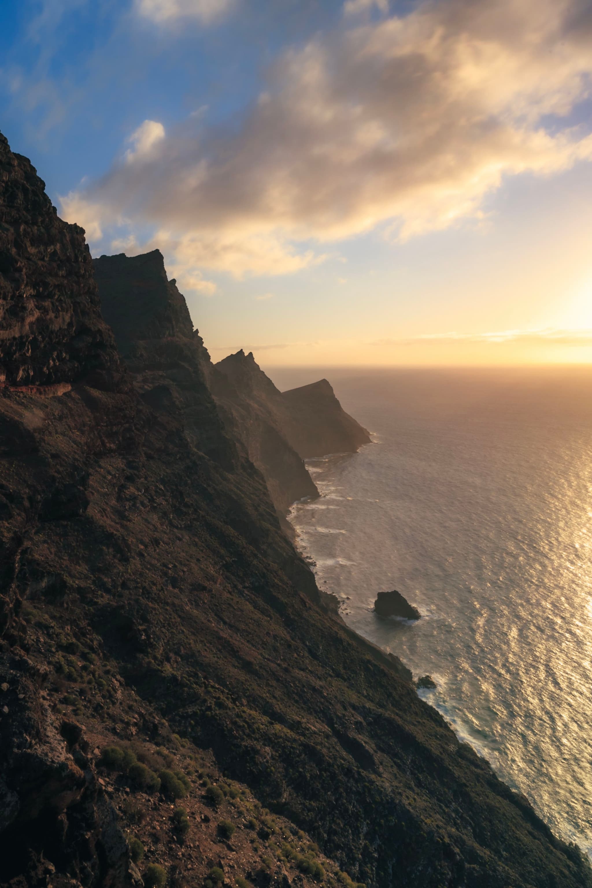 A dramatic coastal cliffside at sunset, with rugged terrain and the ocean stretching into the horizon under a partly cloudy sky