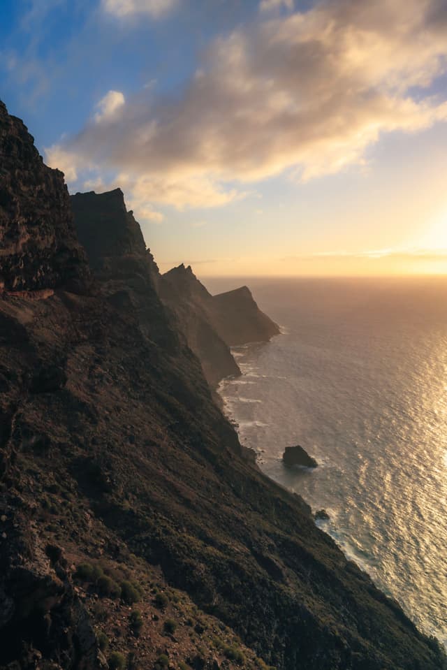 A dramatic coastal cliffside at sunset, with rugged terrain and the ocean stretching into the horizon under a partly cloudy sky