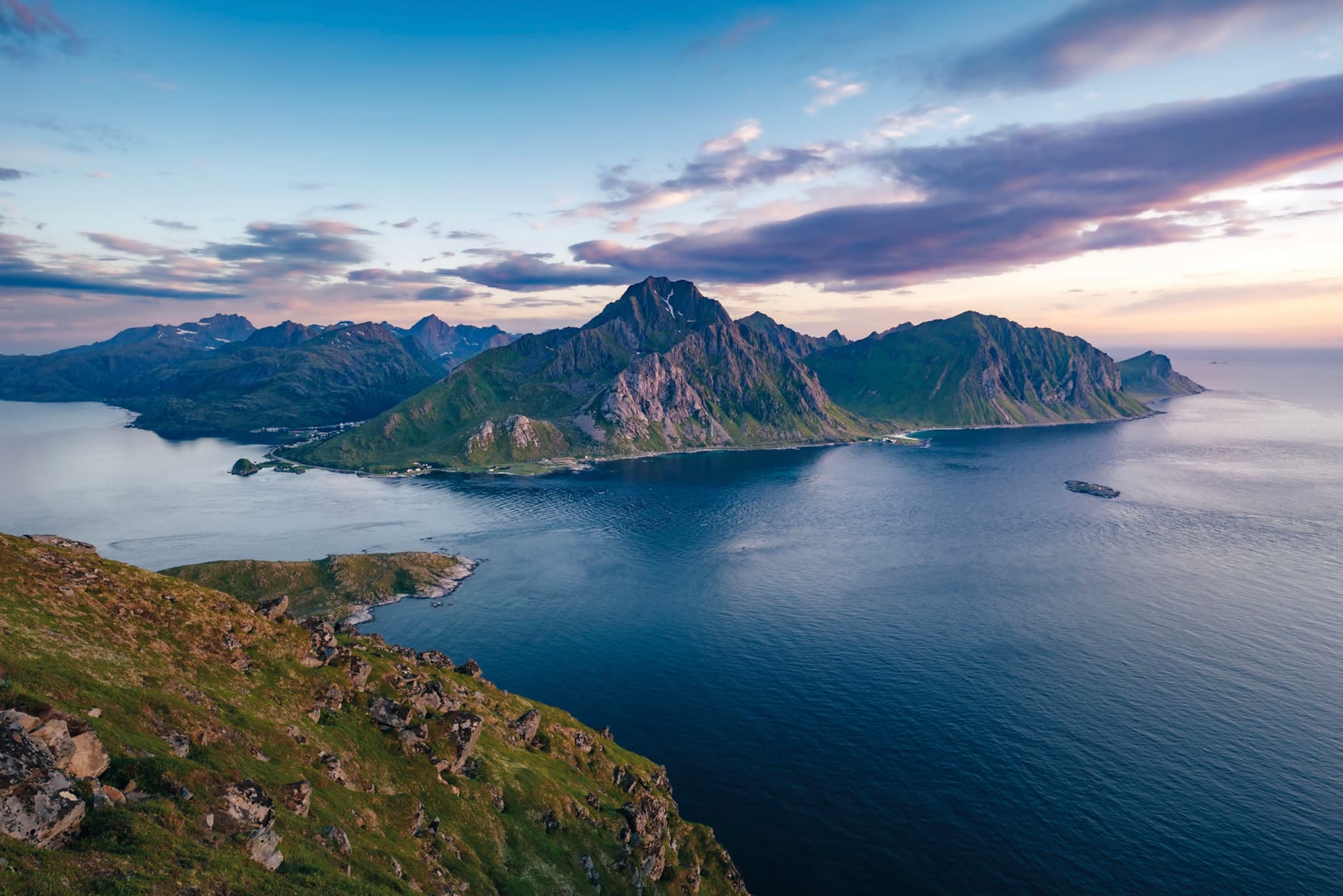 A scenic coastal landscape with rugged mountains, calm blue water, and a partly cloudy sky during sunset