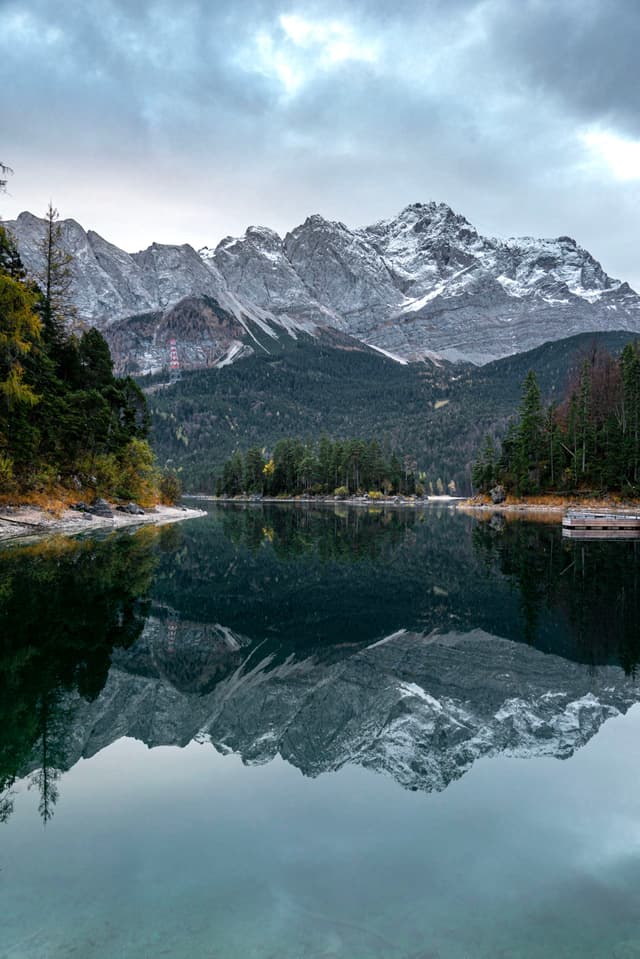 A serene mountain landscape with snow-capped peaks reflected in a calm lake, surrounded by lush forests under a cloudy sky