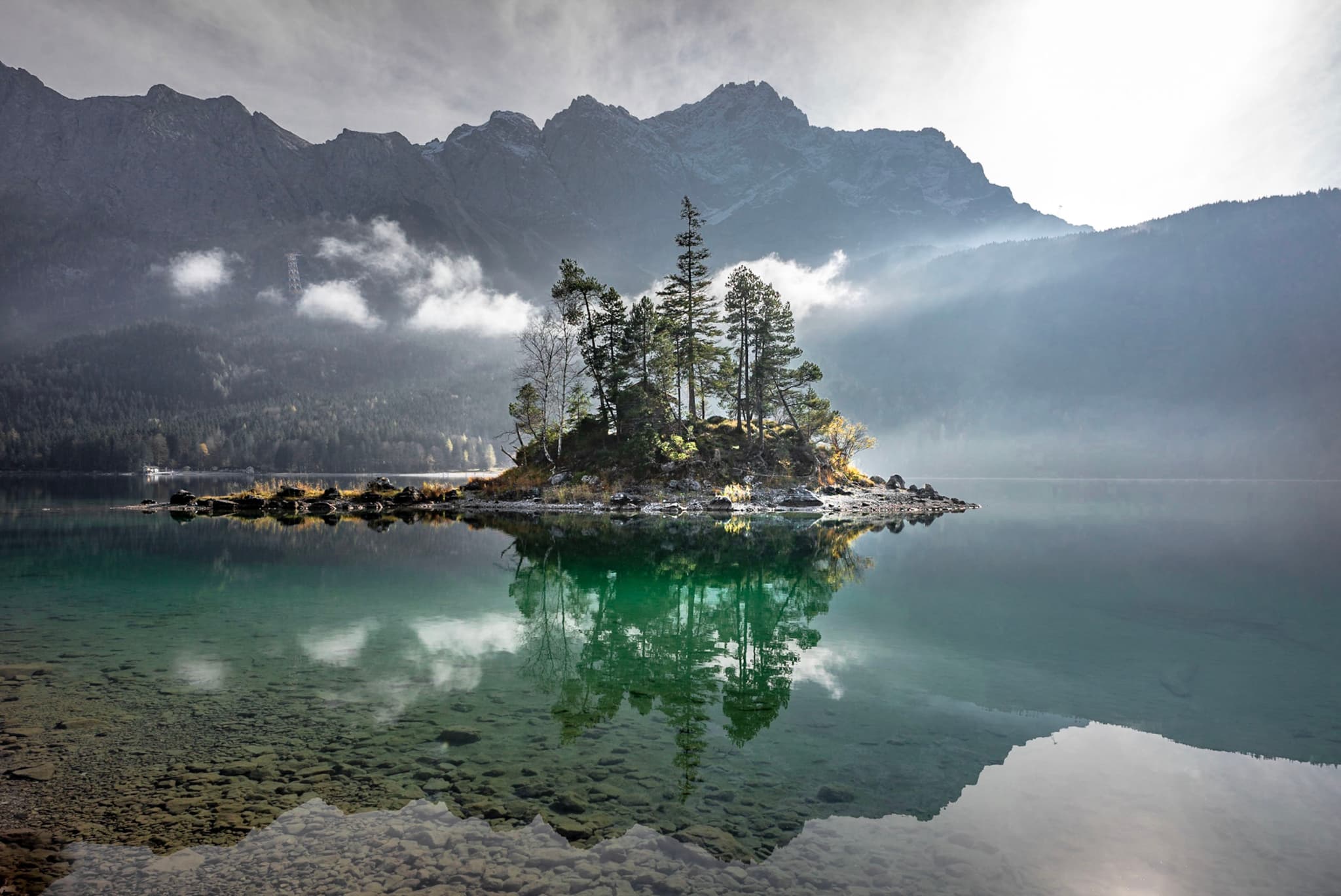 A small, tree-covered island is reflected in a calm lake, surrounded by misty mountains under a bright sky