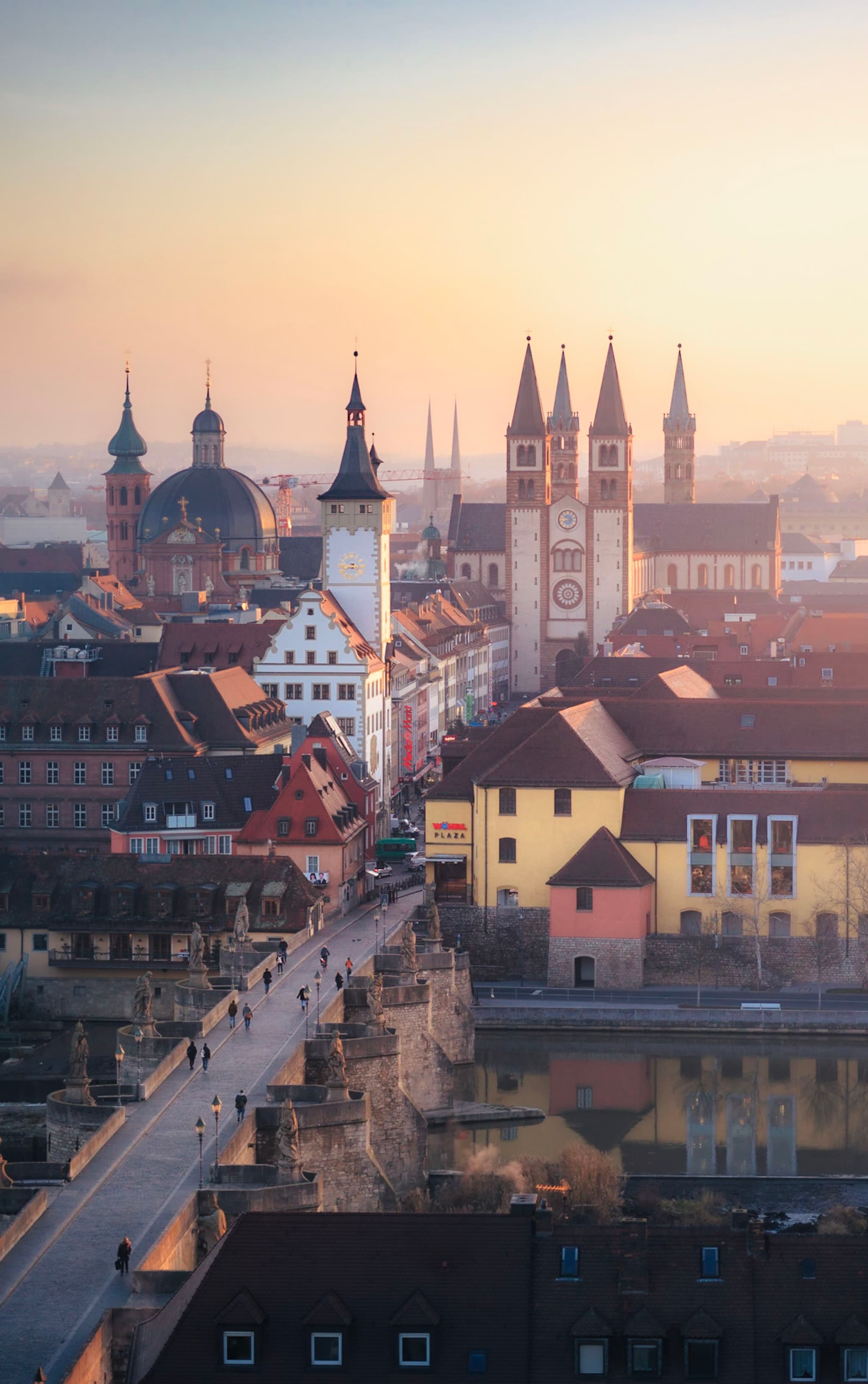 historic European cityscape at sunrise with church towers rising above colorful riverside buildings and a stone bridge filled with pedestrians