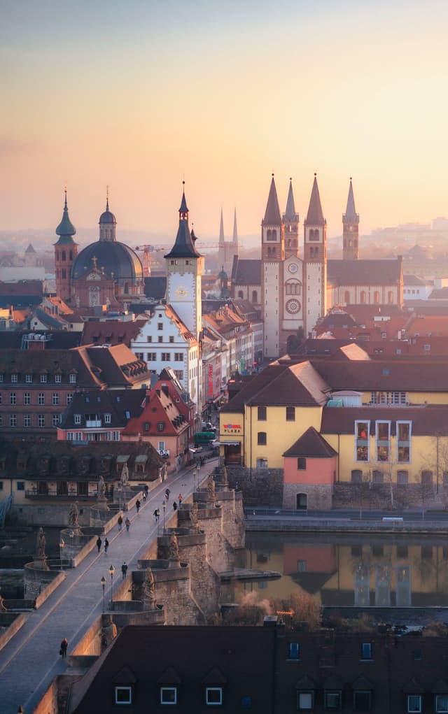 historic European cityscape at sunrise with church towers rising above colorful riverside buildings and a stone bridge filled with pedestrians