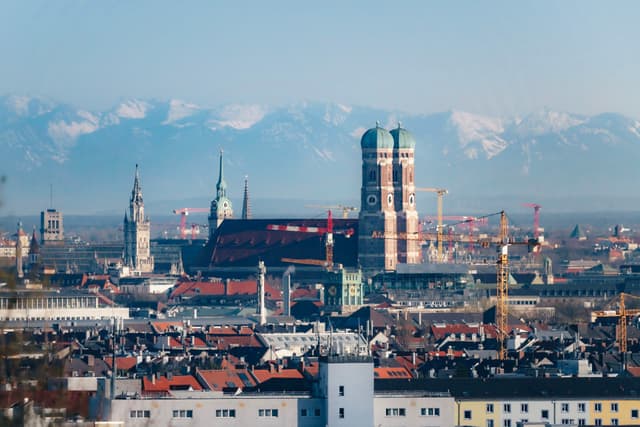 European city skyline with historic church towers and rooftops set against snow-capped alpine mountains in the distance