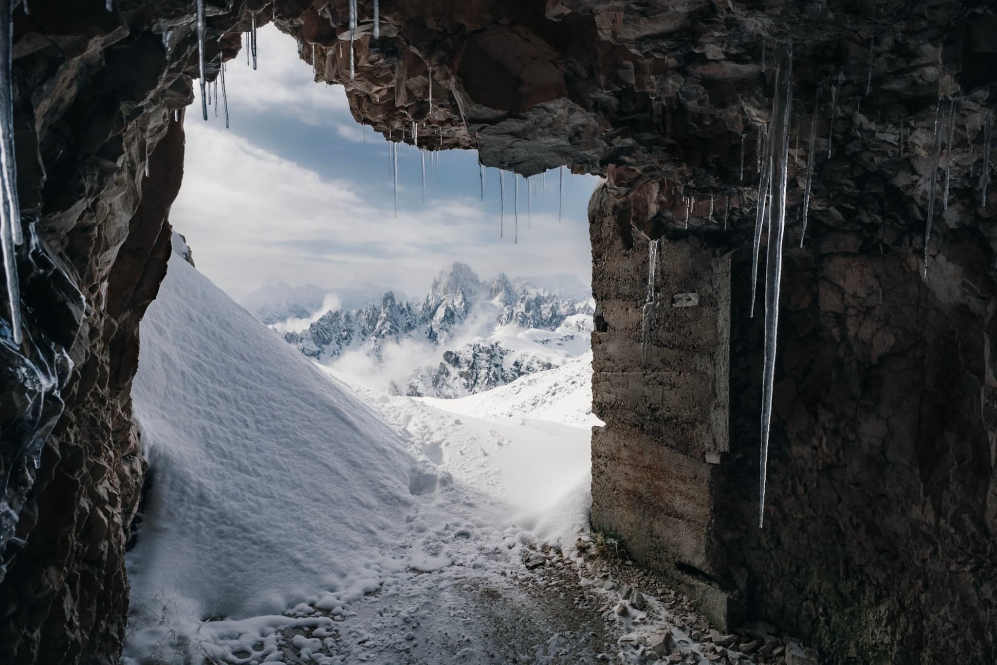 A snowy mountain landscape viewed from inside a rocky cave, with icicles hanging from the entrance