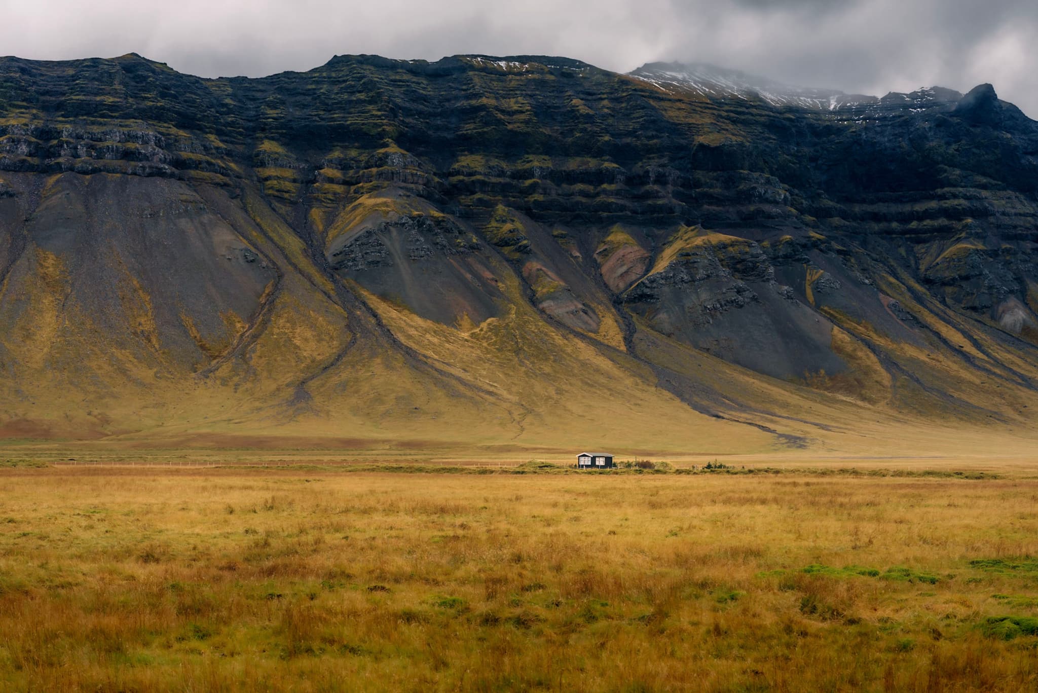 Vast golden valley with a tiny isolated farmstead at the base of steep, layered mountains under low clouds