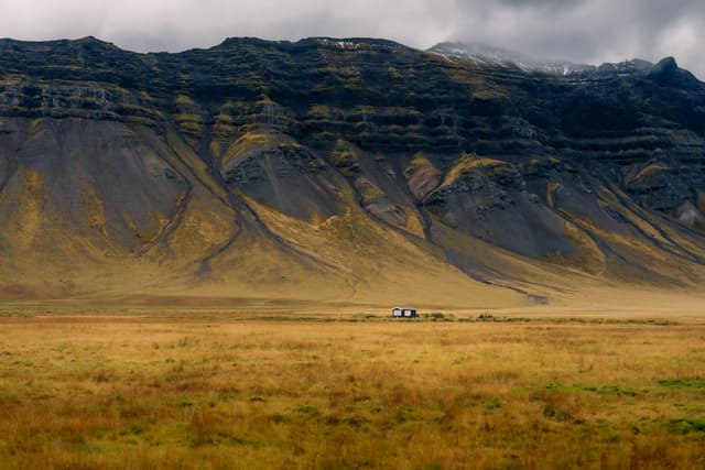 Vast golden valley with a tiny isolated farmstead at the base of steep, layered mountains under low clouds