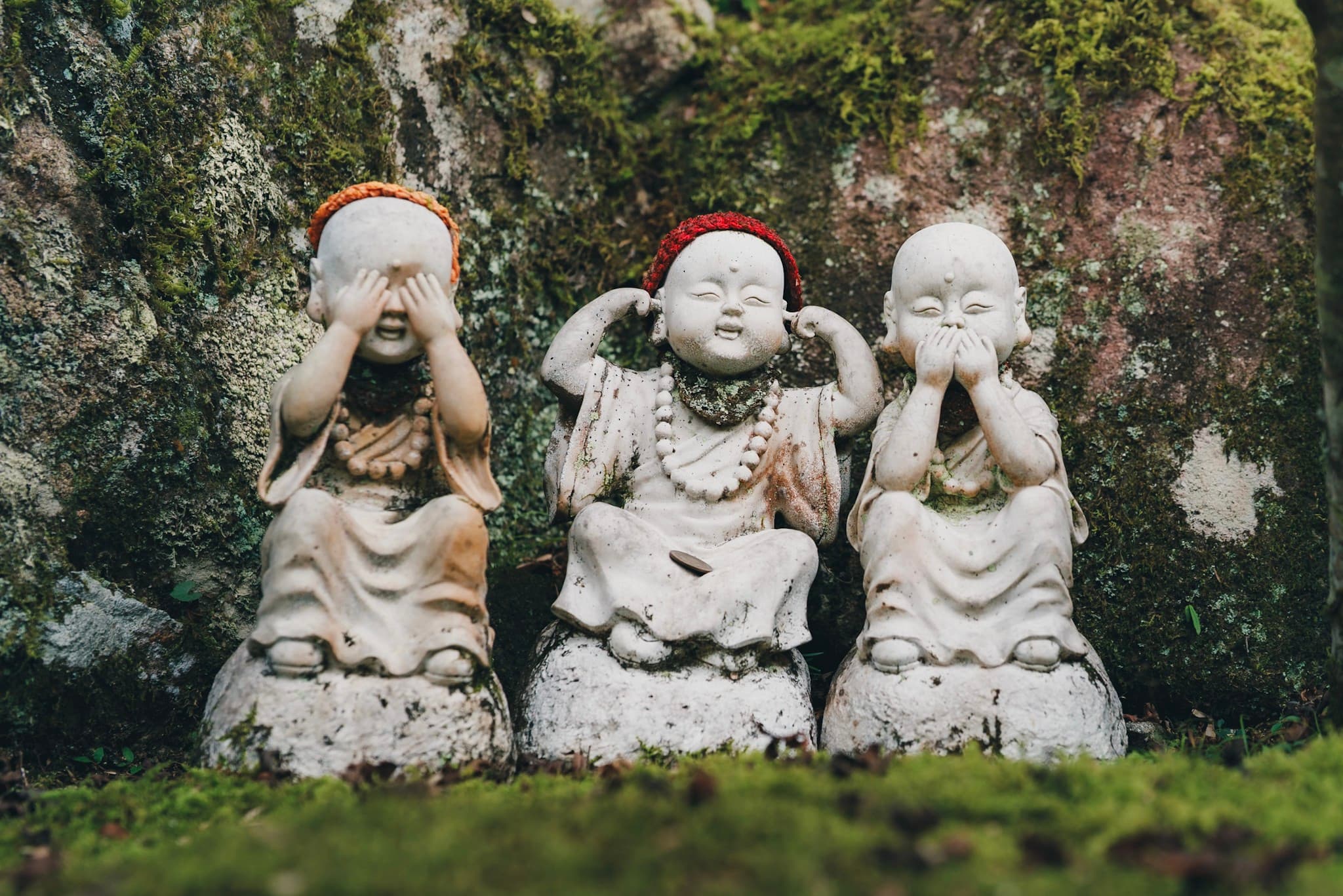 Three small monk statues in the classic see-no-evil, hear-no-evil, speak-no-evil poses against a moss-covered stone background
