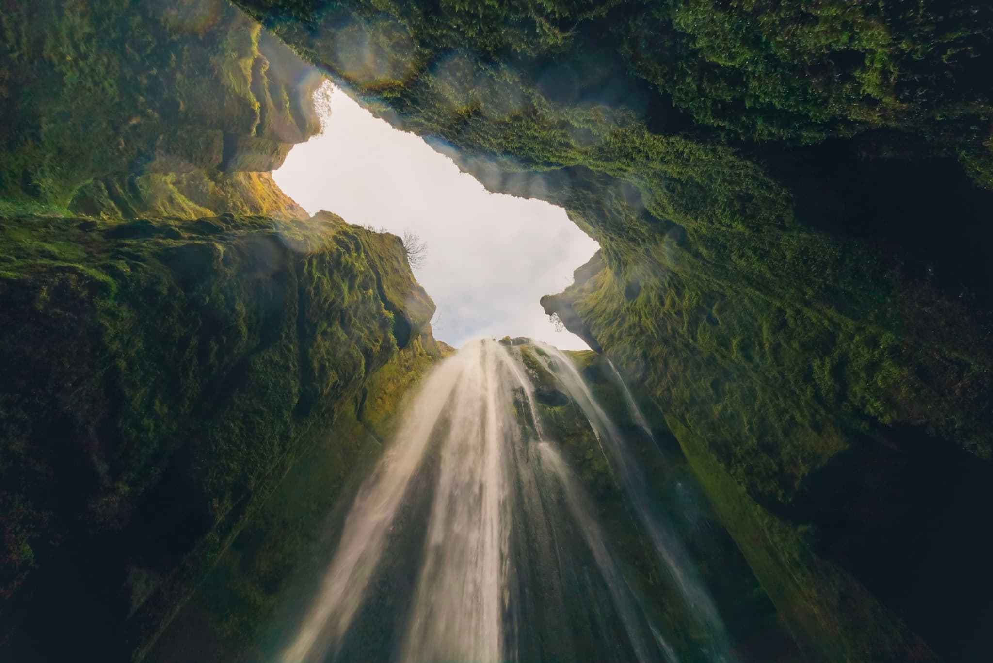 Looking up from the base of a narrow canyon toward a tall waterfall pouring down between moss-covered rock walls into the light above