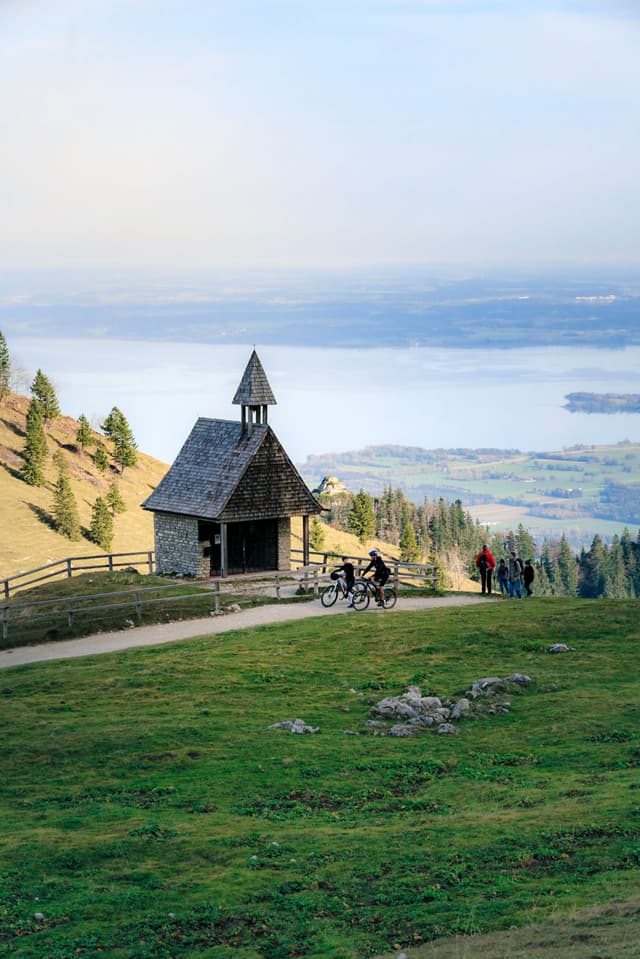 A small chapel with a pointed roof stands on a grassy hillside, overlooking a vast body of water and distant land Two people with bicycles are nearby, and the scene is surrounded by trees and a wooden fence