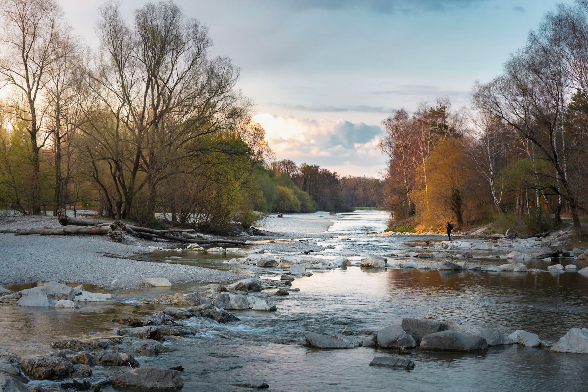 Shallow river flowing over scattered rocks between autumn trees under a soft evening sky with a solitary person walking along the bank